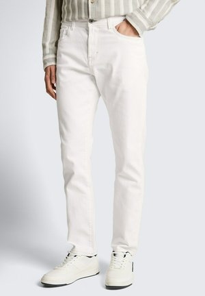 Mężczyzna noszący białe jeansy o prostych nogawkach, białe trampki oraz beżową koszulę w paski z długim rękawem, z jedną ręką w kieszeni.