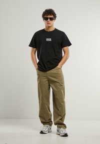 Svart bomull T-skjorte med "USA" logo, kombinert med khaki vid bukse og sølv atletiske sko. Modellen har på seg svarte solbriller, med hendene i lommene.