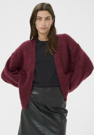 Burgunderfarbener, gestrickter Cardigan mit lockerem Schnitt und offener Vorderseite, aus weichem, strukturiertem Stoff gefertigt. Kombiniert mit einem schwarzen Oberteil und einem Lederrock.