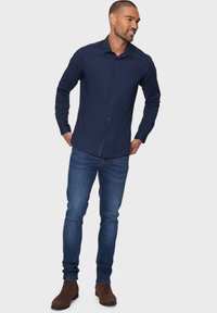 Donkerblauwe langaste mouwblouse met knoopsluiting aan de voorkant, gecombineerd met aansluitende blauwe jeans en bruine enkelboots. Zachte textuur en klassieke snit.