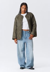 Woman with braided hair wearing an olive quilted jacket, white crop top, wide light blue jeans, black shoes, and rings on her fingers.