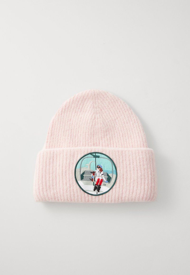 Rozā tamborēta beanie cepure ar ribotu tekstūru, kura vidū ir noapaļots izšūts svešais attēls ar figūru, kas atrodas gondolā kopā ar suni uz zilā fona.