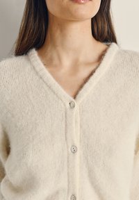 Krémový cardigan vyrobený z měkkého, nadýchaného materiálu. Obsahuje výstřih do V a šest knoflíků s jemným leskem. Texturovaný pletený vzor.