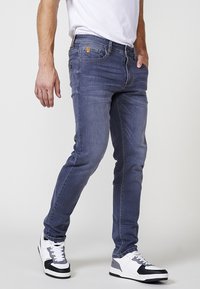Jeans de mezclilla en un lavado azul medio, de corte slim, con un diseño estándar de cinco bolsillos y detalles de metal dorado. Combinados con zapatillas blancas y negras.