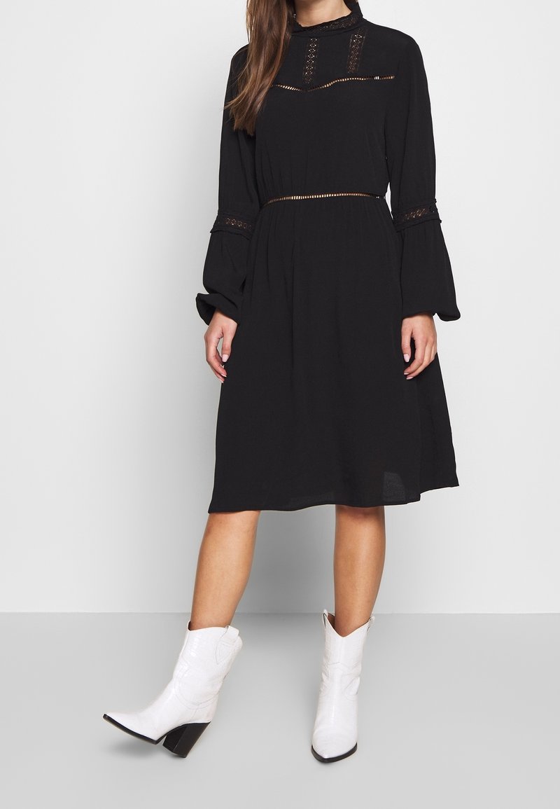 Femme portant une robe mi-longue noire à manches longues avec des détails en dentelle, associée à des bottines blanches à bout pointu et talons carrés.