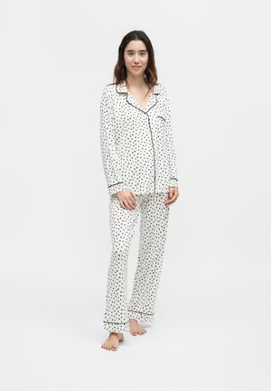 Νεαρή γυναίκα στέκεται ξυπόλητη, φορώντας λευκές πιτζάμες με μακριά μανίκια και κουμπιά, με μικρά μαύρα σχέδια καρδιάς και μαύρο περίγραμμα.