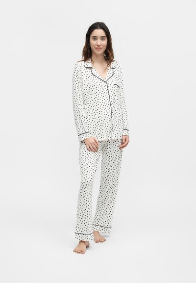 Jeune femme debout pieds nus portant un pyjama blanc à manches longues boutonné avec de petits motifs de cœurs noirs et un passepoil noir.