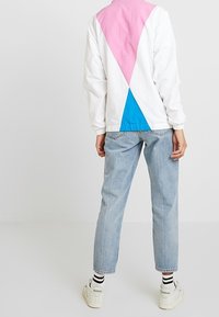 Biała kurtka przeciwwiatrowa z różowymi i niebieskimiGeometrycznymi wzorami na plecach, zestawiona z jasnoniebieskimi dżinsami i białymi trampkami w czarne paski.