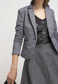 Grijs tweed blazer met ingeknipte revers, zwarte accenten en aansluitende mouwen over een zwart graphic shirt en een grijze flared rok.