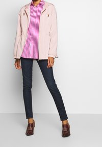 Ljust rosa zip-up jacka med huva, matchad med en randig skjorta i rosa och vitt, mörkblå jeans och bruna skor med accentdetaljer.
