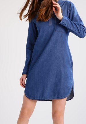 Mujer con vestido de mezclilla azul de manga larga, con bolsillo delantero y bajo redondeado, posando con el cabello cubriendo parcialmente el rostro.