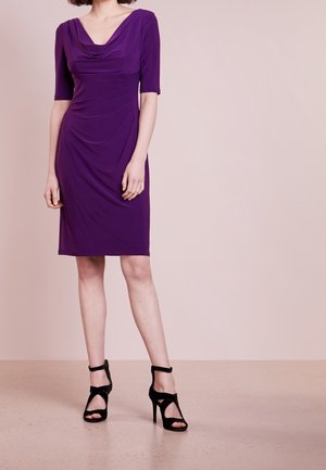 Woman wearing a purple knee-length dress with three-quarter sleeves and black high-heeled sandals against a plain beige background.