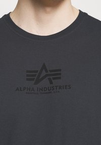 Tmavě šedé tričko s černým logem obsahujícím geometrické tvary a text "ALPHA INDUSTRIES" a "KNOXVILLE, TENNESSEE, U.S.A."