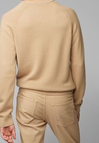 Béžový pletený svetr s žebrovaným límcem a manžetami, kombinovaný s béžovými kalhotami, které mají dvojité zadní kapsy. Texturovaná látka s jemným povrchem.