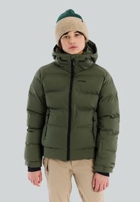 Jongere met een beige en groene gebreide muts, een groene gewatteerde jas met capuchon en beige broek, staand met een neutrale uitdrukking.