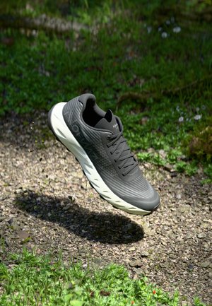 Gray athletic shoe with textured mesh upper and lace closure, featuring a white midsole and cushioned outsole, suspended above gravel and grass.
