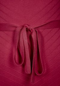 Neulemekko syvän burgundinpunaisena, jossa on teksturoitu diagonaalinen kuvio. Vyötäröllä kangasnauha, joka korostaa sileää ja pehmeää pintaa.