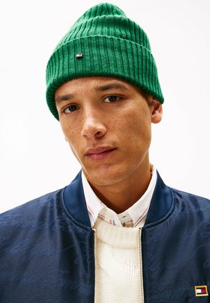 Berretto a maglia verde con texture a coste e piccolo logo, indossato con una giacca bomber blu, un maglione bianco e una camicia con colletto sotto.