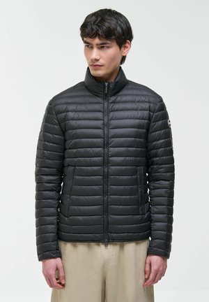 Giovane uomo che indossa una giacca trapuntata nera con cerniera e pantaloni beige, in piedi davanti a uno sfondo grigio chiaro uniforme.