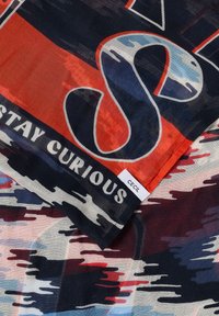 Tissu avec un motif camouflage audacieux en rouge, bleu et noir, un grand "S" stylisé, le texte "Stay Curious" et une petite étiquette "CECIL".