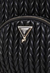 Sac à main en cuir synthétique tissé noir avec un logo triangulaire Guess et des accents dorés. Détail de zip sur le dessus pour une fermeture sécurisée.