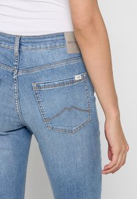 Jean en denim bleu délavé avec une coupe ajustée. Présente une couture décorative marron sur les poches arrière et une étiquette de marque grise à la taille.