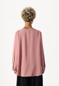 Langermet, løs skjorte i myk rosa stoff. Har rund hals og bakfold for økt bevegelse. Minimalistisk design.