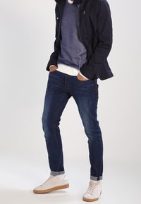 Námořnická bunda na zip přes modrý svetr s bílými manžetami, doplněná o úzké tmavé džíny a bílé tenisky s béžovými podrážkami.