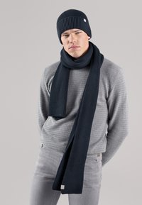 Navy ribbed knit beanie and scarf set, featuring a soft texture and neat edges, paired with a light grey patterned sweater.