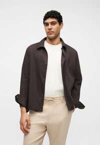 Άνδρας με ανοικτό σκούρο καφέ σακάκι πάνω από άσπρο πουκάμισο και μπεζ παντελόνι με κορδόνι, στέκεται μπροστά σε απλό φωτεινό φόντο.