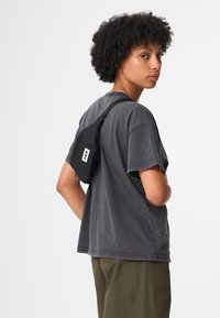 Schwarze Umhängetasche aus Stoff mit einem seitlichen Reißverschluss und einem Logopatch. Wird auf dem Rücken getragen, kombiniert mit einem grauen T-Shirt und olivgrünen Hosen.