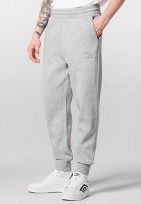 Pantalons de survêtement gris avec une taille élastique, des poches latérales et un logo brodé, associés à des baskets blanches avec des accents noirs.