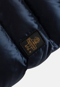 Granatowa puchowa kurtka o gładkim satynowym wykończeniu, z prostokątną czarną metką z ilustracją złotego psa i nazwą marki.