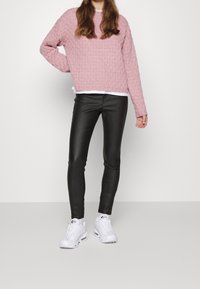 Kvinna som bär en rosa stickad tröja, svarta skinnbyxor och vita sneakers, stående mot en enkel vit bakgrund.