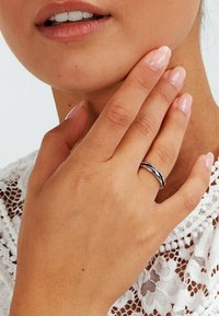 Närbild av en hand med polerade naglar som vilar på ett hakan, bärande en enkel silverring, mot en vit spetsklädsel.