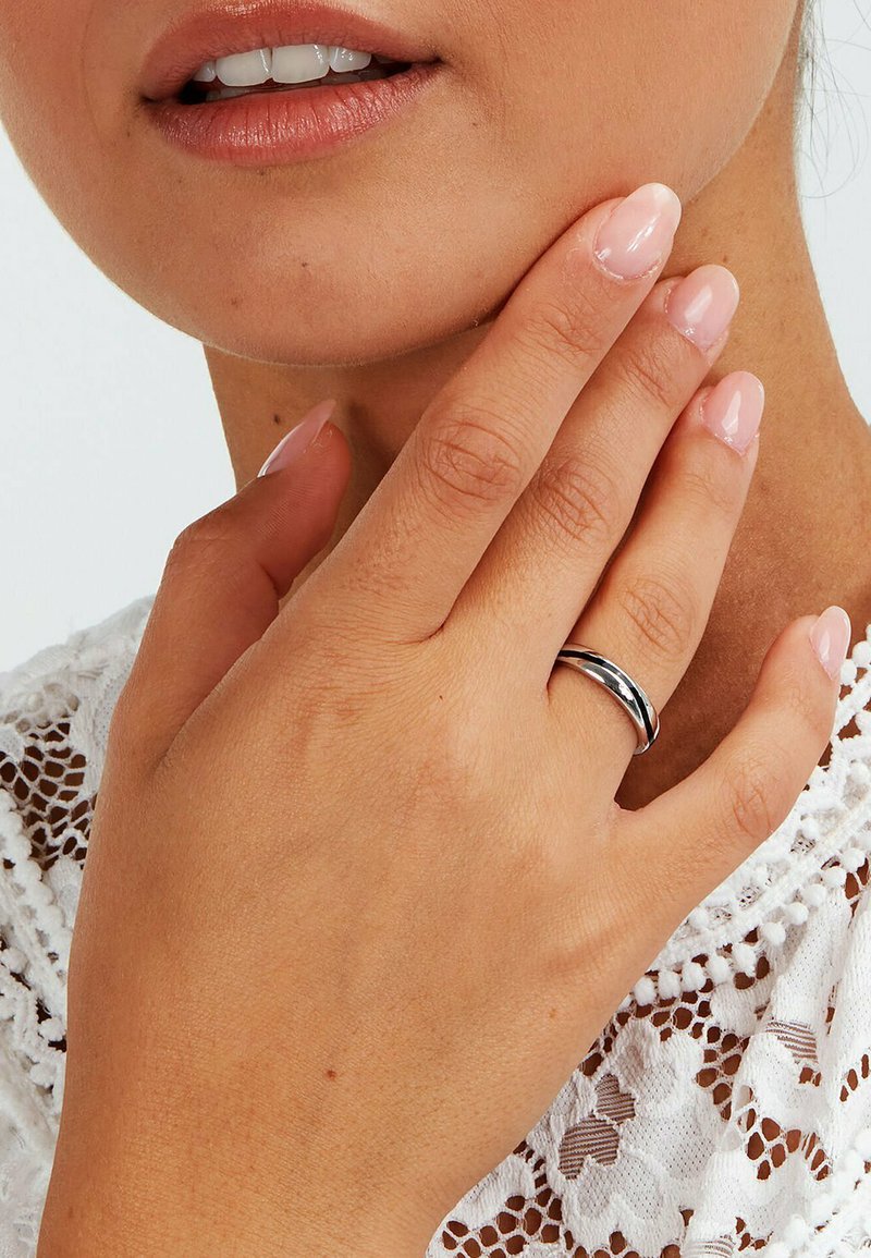 Närbild av en hand med polerade naglar som vilar på ett hakan, bärande en enkel silverring, mot en vit spetsklädsel.