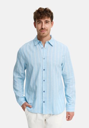 Mand iført en lyseblå, langærmet, vertikalt striped skjorte med hvide bukser mod en ensfarvet baggrund.