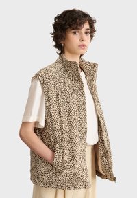 Beige kviltad väst med svart leopardtryck, med hög krage, dragkedjestängning och sidofickor, bärs över en vit skjorta.