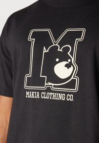 Černé bavlněné tričko s velkým bílým obrysovým písmem "M" a grafikou medvěda, s nápisem "MAKIA CLOTHING CO." tištěným níže v bílé barvě.
