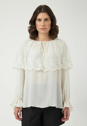 Kvinde med medium brunt bølget hår bærer hvid bluse med blondecapelet og lange flæseærmer, parret med sorte bukser, stående frontalt.