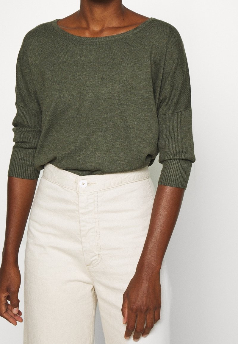 Olivgrön stickad tröja med bred halsringning och ribbade ärmslut, tillsammans med ljusbeige högmidjade byxor med knappstängning.