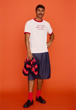 Mann mit Schnurrbart, der ein weißes T-Shirt mit rotem Besatz und Text, marineblaue Shorts, knallrote Socken, schwarze Schuhe trägt und eine gestreifte Jacke hält, steht vor einem orangenen Hintergrund.