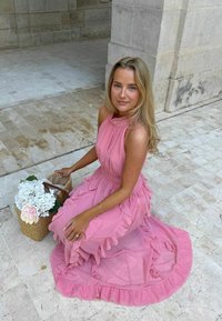 Jeune femme en robe rose fluide assise sur un sol en pierre près d’un panier de fleurs blanches et roses, avec des murs en pierre à l’arrière-plan.