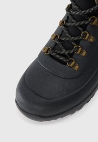 Bota de caminhada preta com biqueira reforçada, sola de borracha texturizada e atacadores pretos cruzados com ganchos de metal em fundo branco.