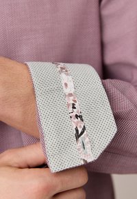 Primo piano di una mano che sistema il polsino di una camicia rosa con trama, con un risvolto bianco a contrasto caratterizzato da una striscia con occhielli dal motivo floreale.