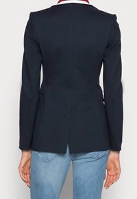 Granatowa marynarka z długimi rękawami, o strukturalnym kroju i rozcięciu z tyłu. Z kontrastującym bordowym kołnierzem z białą lamówką. Nosi się ją na jasnoniebieskie dżinsy.