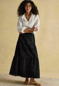 Biała bluzka z ażurowym wzorem i falbanianym kołnierzem, zestawiona z czarną, warstwową, bawełnianą maksi spódnicą ozdobioną okrągłym haftem oraz żółtymi sandałami.