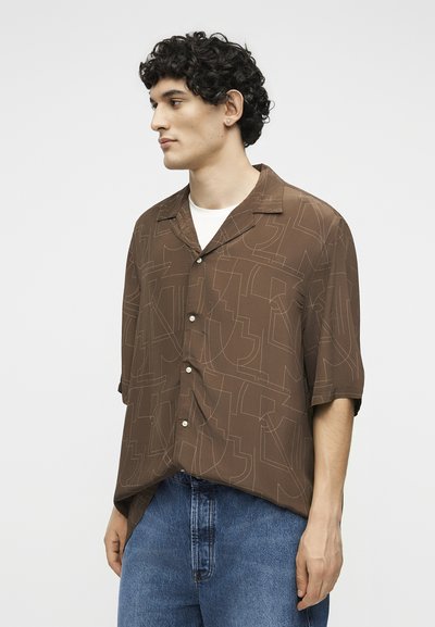 Νέος άντρας με σγουρά μαλλιά, φορώντας ένα καφέ κοντομάνικο πουκάμισο με αφηρημένα σχέδια και μπλε τζιν, κοιτάζοντας προς τα αριστερά.