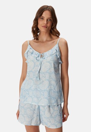 Vrouw draagt lichtblauw paisleyprint mouwloos topje met ruches en bijpassende korte broek tegen een effen achtergrond.
