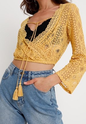 Woman wearing a yellow lace long-sleeve crop top over a black bralette paired with high-waisted blue jeans, hand in pocket.
